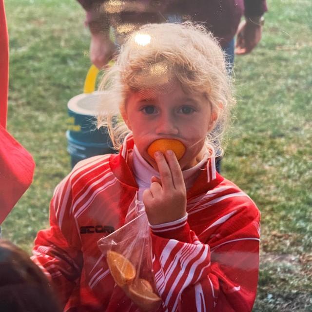 A young girl in a red soccer uniform eating an orange slice.